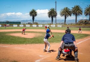 Image from behind home plate of a Little League baseball game. Ump and catcher are ready as right-handed batter stands in the box. Pitcher is on the mound and outfielders are at the ready.