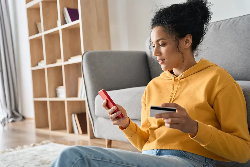 Young adult African American female consumer holding credit card and smartphone sitting on floor at home doing online banking transaction.