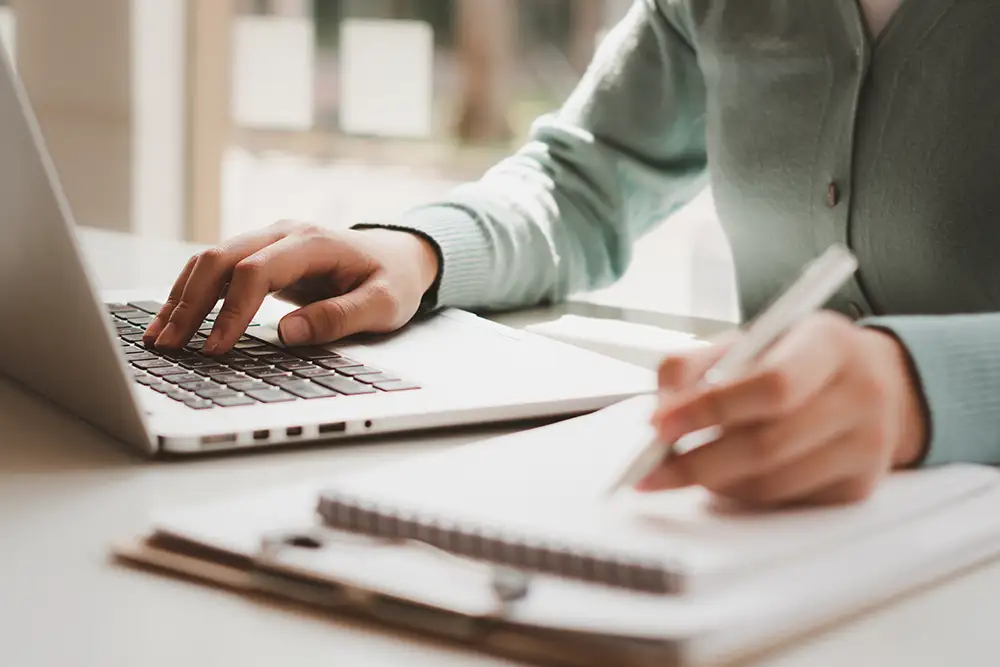 Business women work on computers and write on notepad with pen to calculate financial statements within the office.